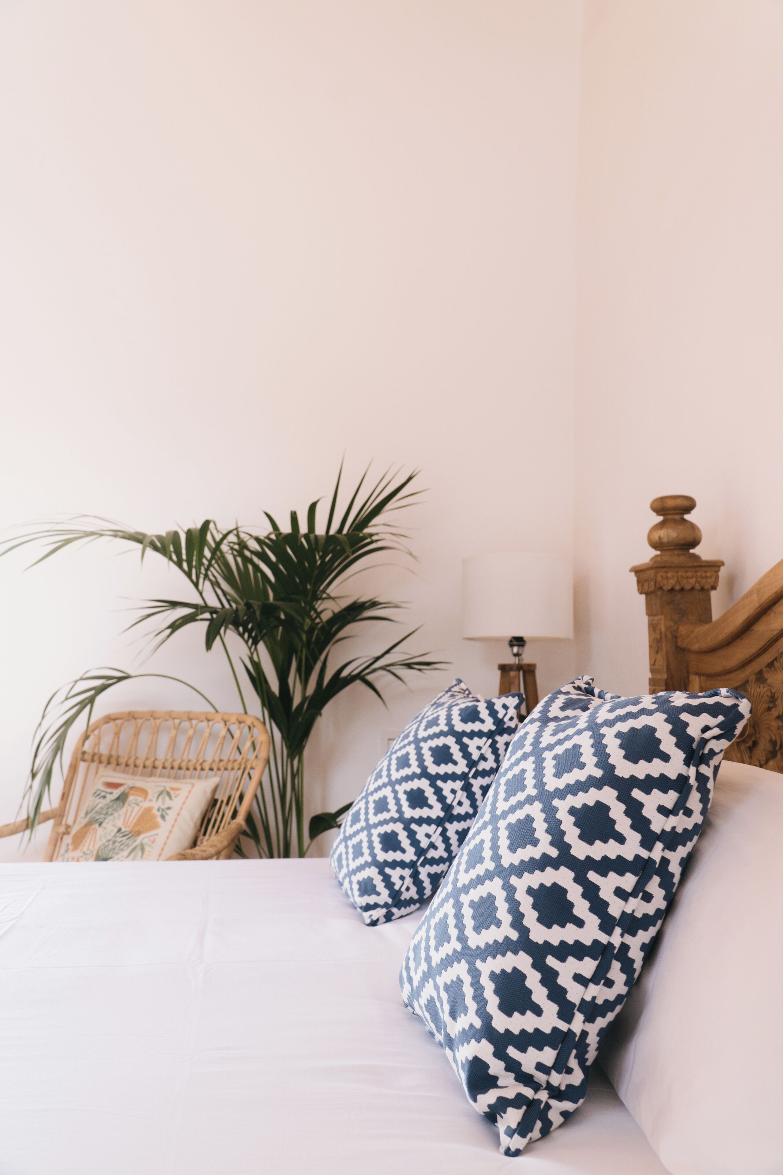 Una cama con ropa de cama blanca y dos almohadas estampadas, un cabecero de madera, una lámpara y una planta en maceta.