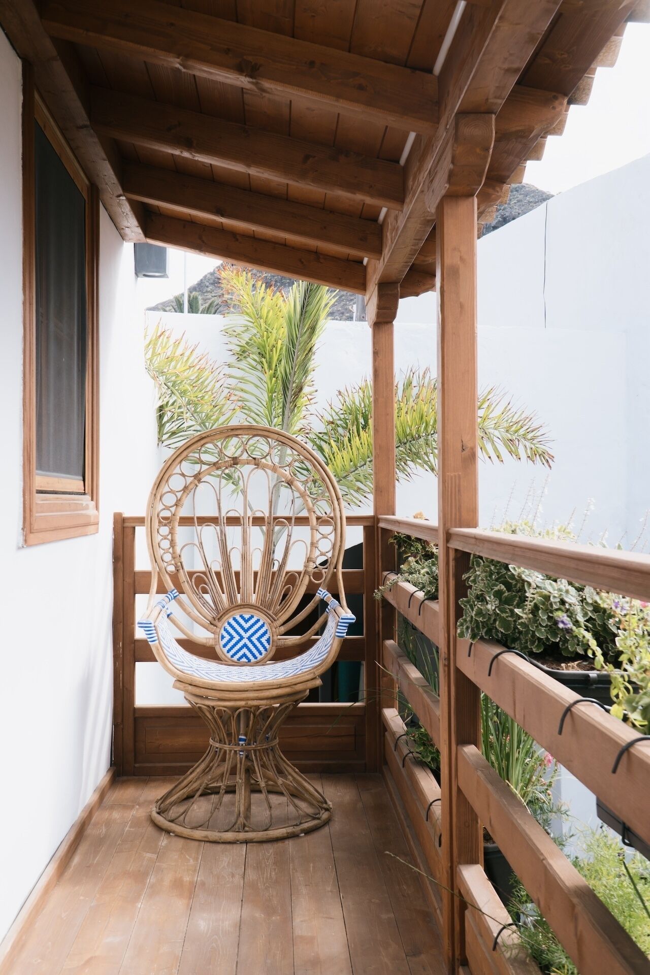 Een houten balkon met een rieten stoel en een tafel, omringd door houten leuningen en plantenbakken.