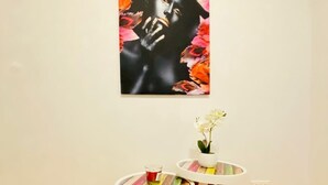 A room with a wooden floor, a white wall, and a painting of a woman with flowers. There are two small tables with colorful wooden tops and a white vase with flowers on one of them. - Sweet 3BR Apt in a Quiet Neighborhood (Vienna)