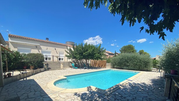 A swimming pool with stone tiles, surrounded by a garden with potted plants and a building with a tiled roof.