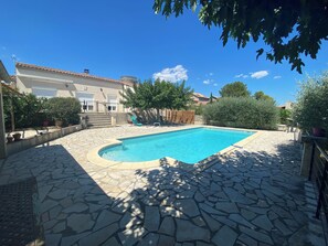 A swimming pool with stone tiles, surrounded by a garden with potted plants and a building with a tiled roof. - Villa in the south (Lecques)