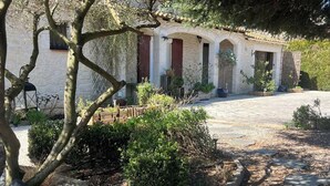 Un edificio de piedra con techo de tejas, camino de grava y jardín con diversas plantas y una estatua.