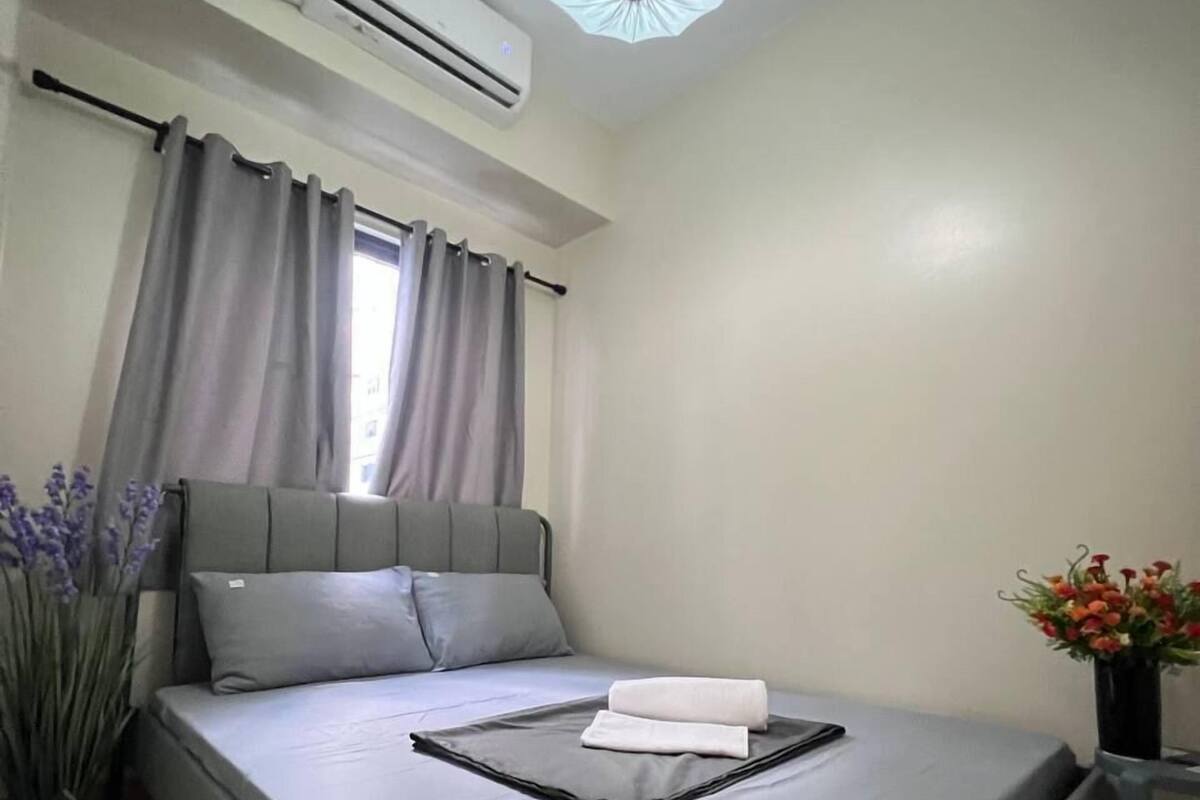 A neatly made bed with a gray mattress and pillows, an air conditioner on the wall, a window with curtains, and a vase with flowers on a nightstand.