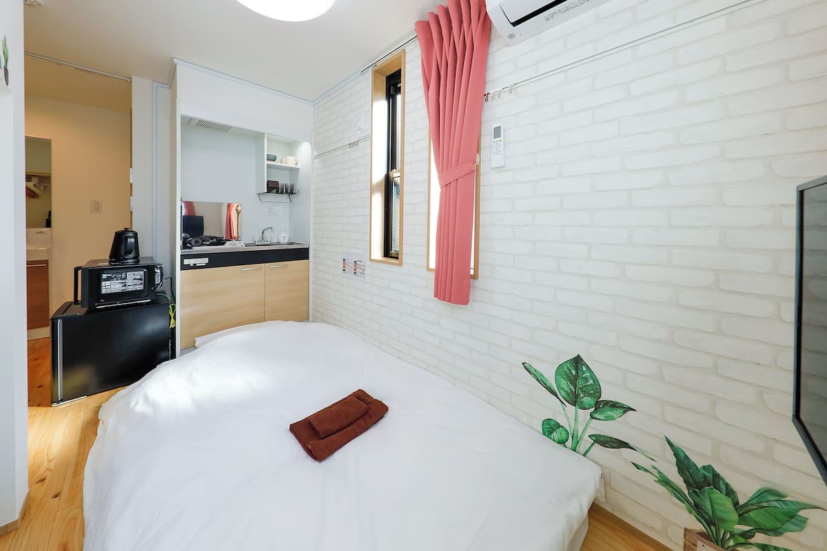 A hotel room with a bed, a small desk with a coffee maker, a mirror, a window with pink curtains, and a wall-mounted TV.