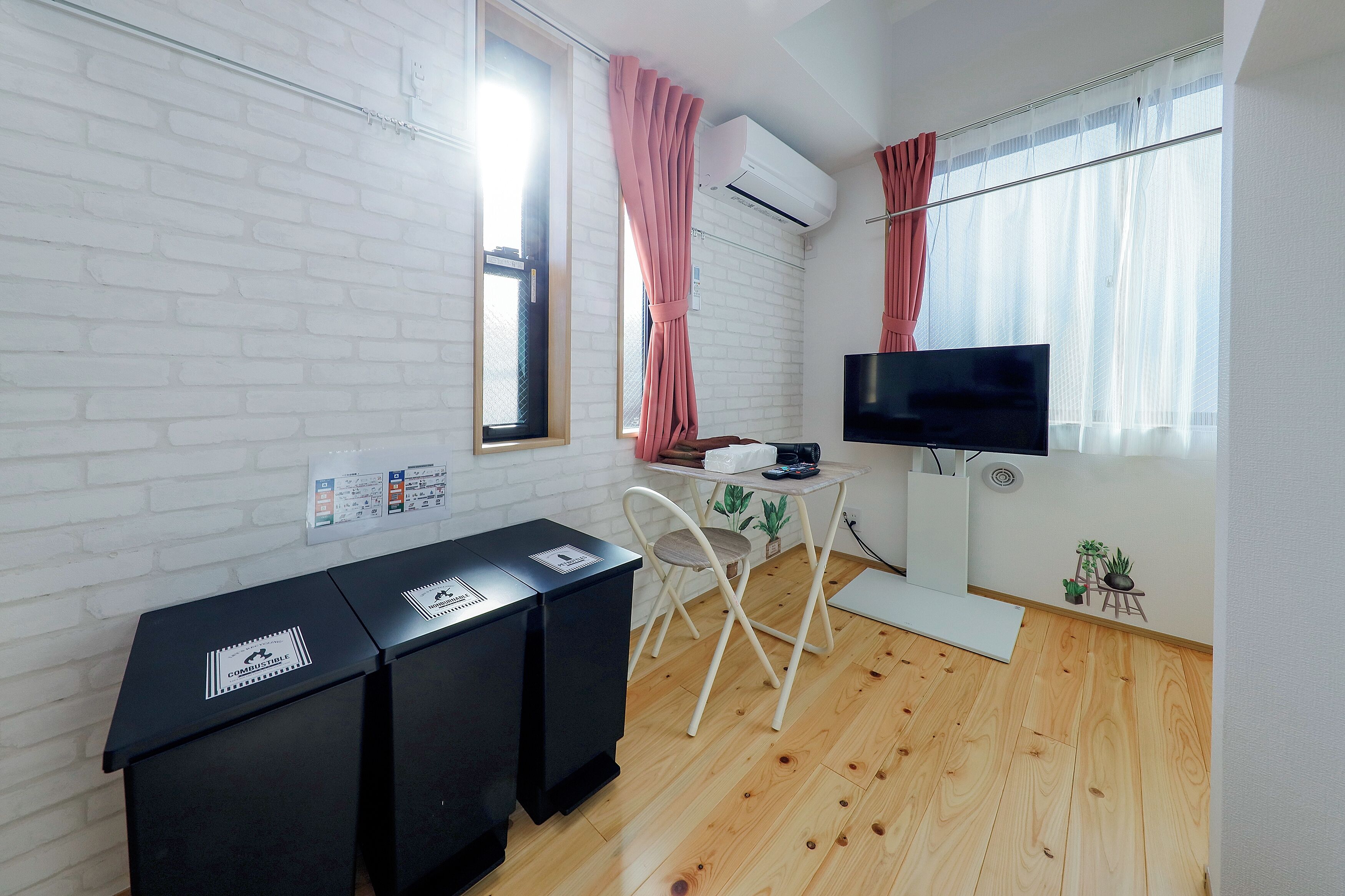 Une pièce de taille modeste avec un mur en briques blanches, un plancher en bois, un bureau avec un ordinateur, une télévision et une fenêtre avec des rideaux.