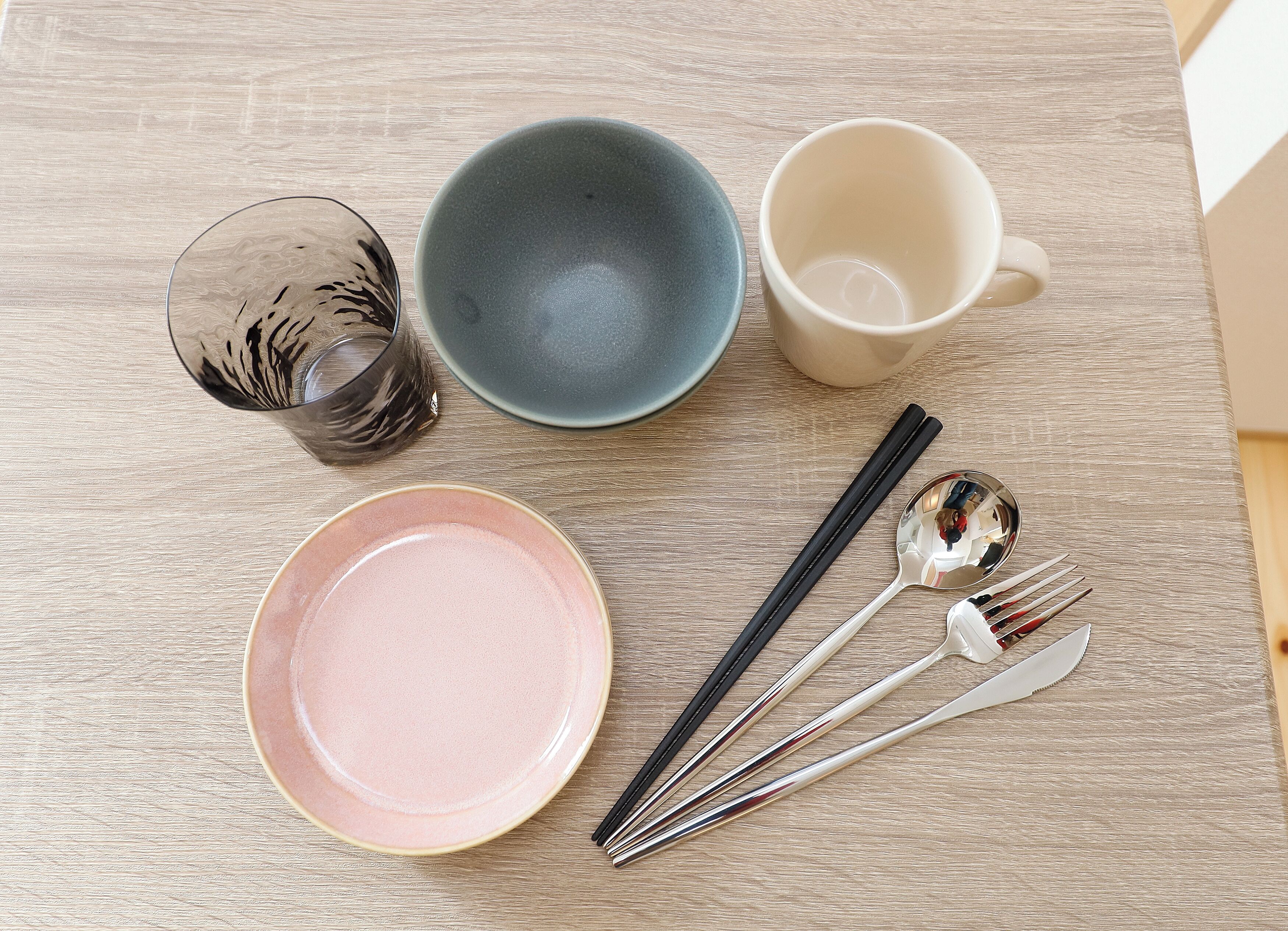Une table avec un verre, un bol bleu, une tasse blanche, une assiette rose, des baguettes et une cuillère.