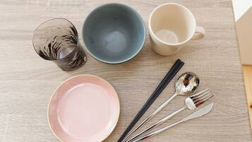 A table with a glass, a blue bowl, a white mug, a pink plate, chopsticks, and a spoon.