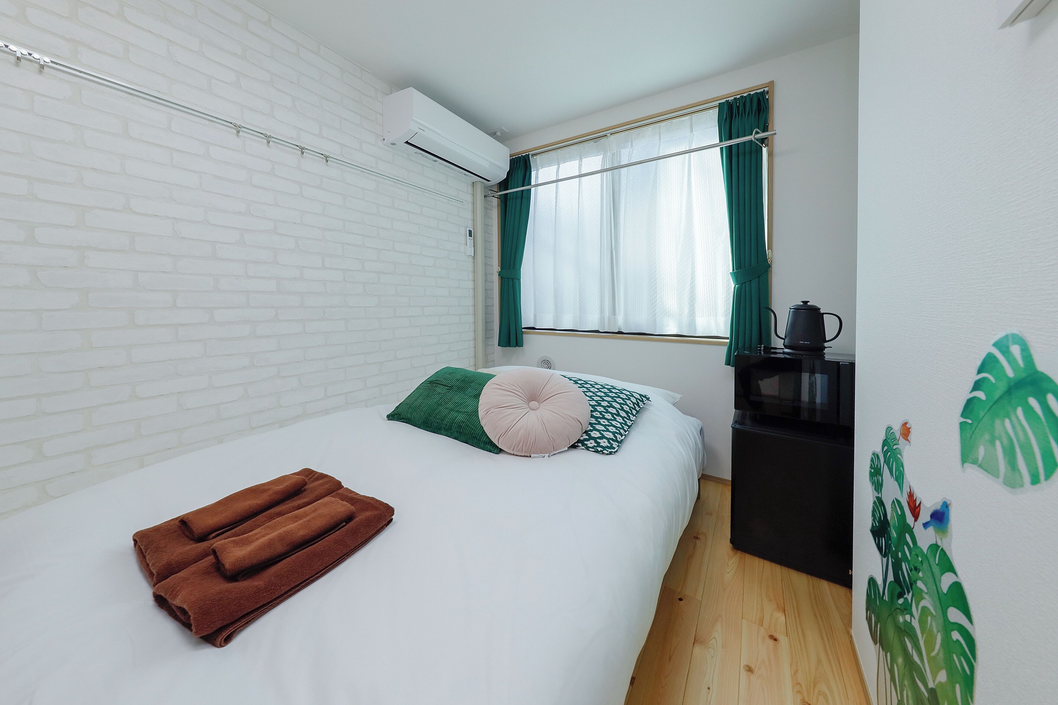 A bedroom with a white brick wall, a bed with a green pillow and a brown towel, a window with green curtains, and a black cabinet with a kettle.