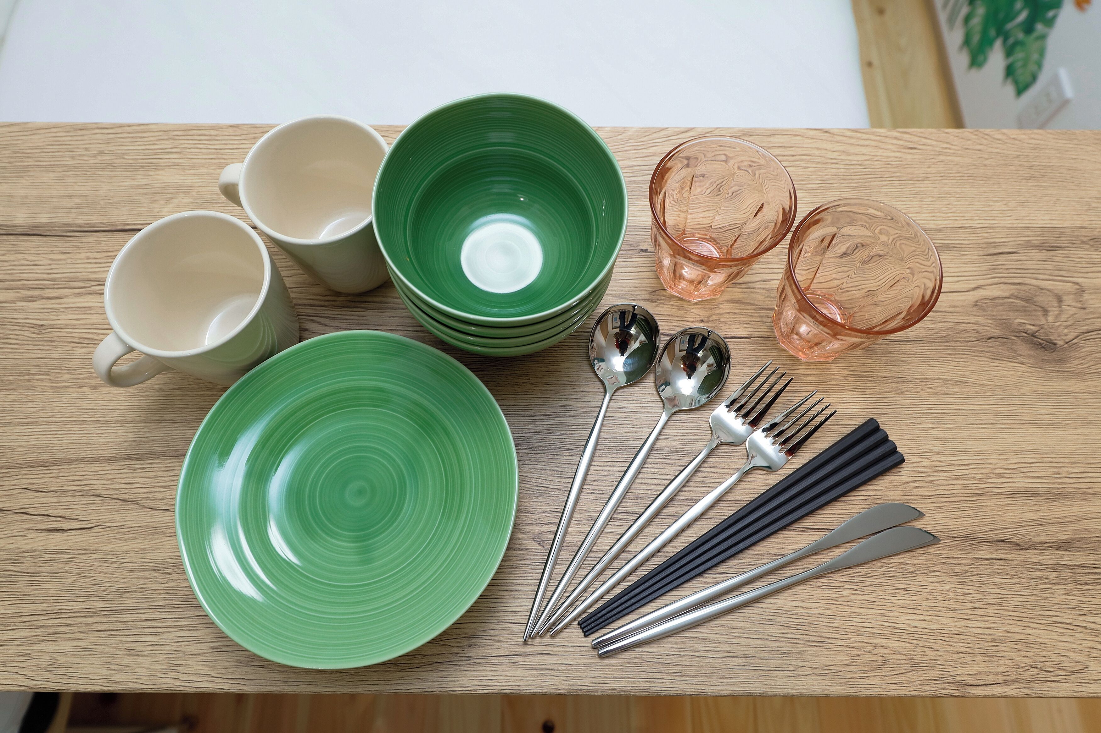 A wooden table with various dishes and utensils, including green plates, white cups, and pink glasses.