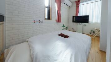 A hotel room with a white brick wall, a flat-screen TV mounted on the wall, a bed with white bedding, and a small table with a plant.