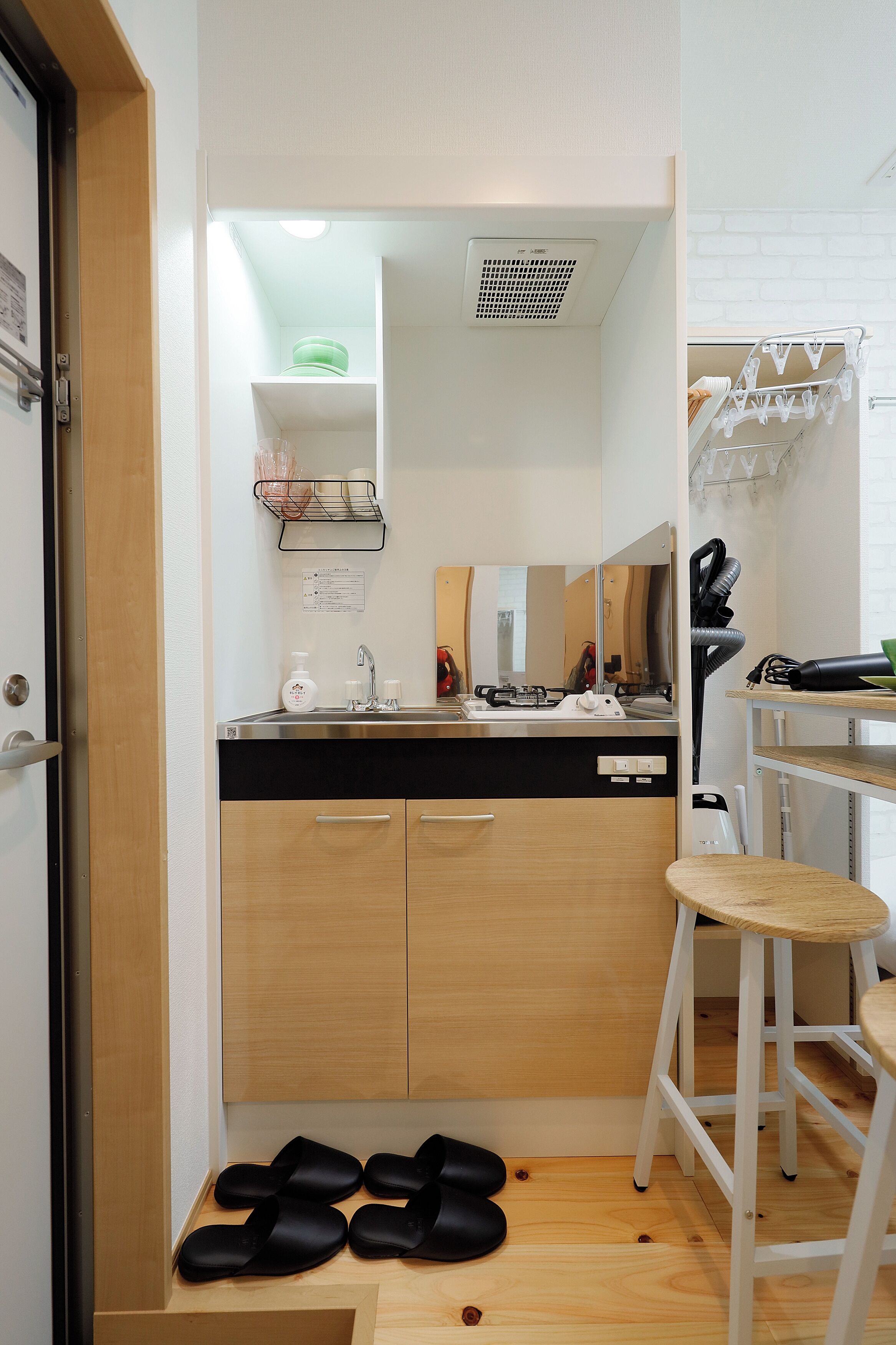 A compact kitchenette with a sink, countertop, and wooden cabinets. There are black slippers on the floor and a white stool near the sink.
