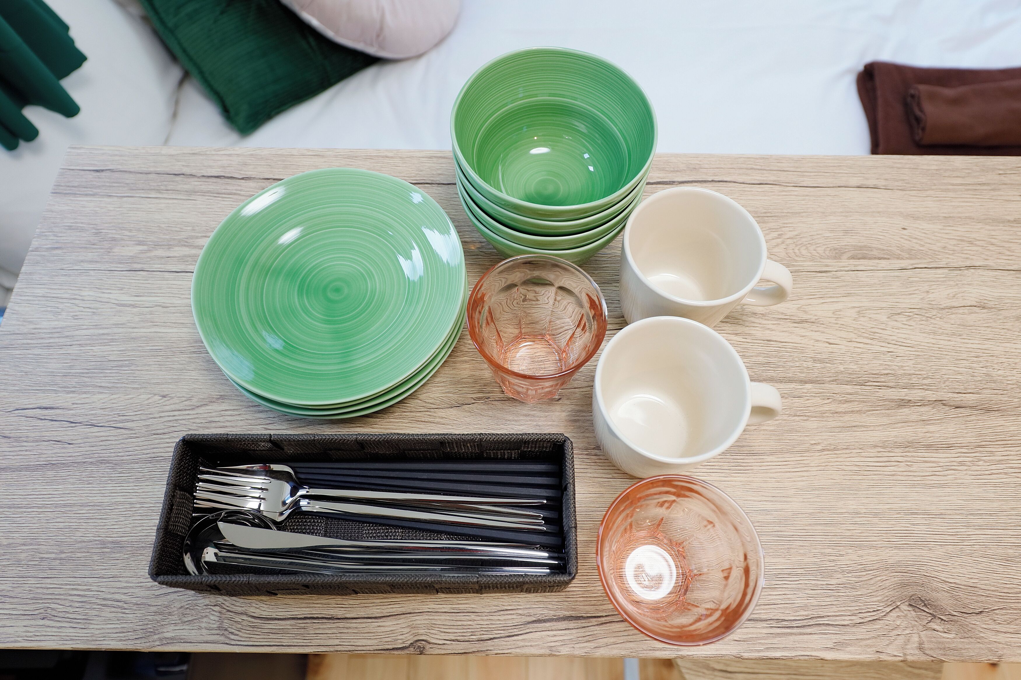 A wooden table with green plates, a glass, a white mug, and a tray with cutlery.