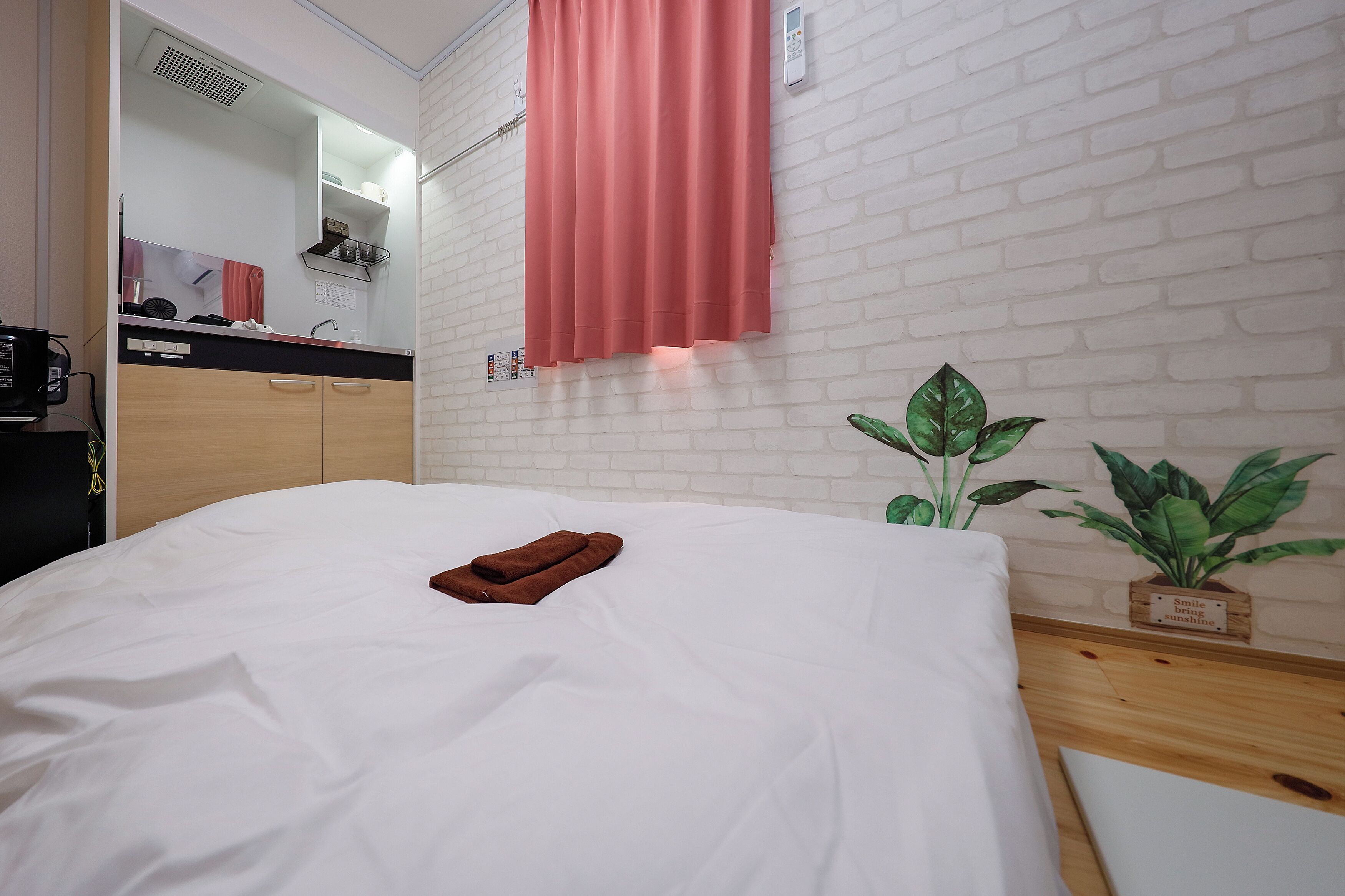 Een netjes opgemaakt bed met een bruine handdoek, een witte bakstenen muur, een rood gordijn en een kleine potplant.