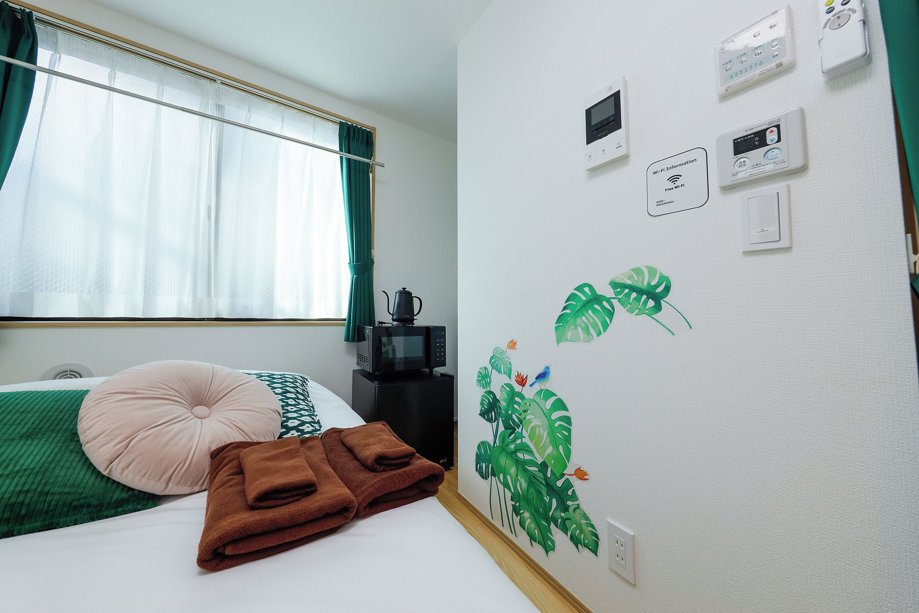 Een hotelkamer met een bed, een kussen met een groen-wit patroon, een bruine handdoek, een groen gordijn, een wandthermostaat en een Wi-Fi sign.