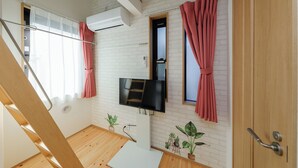 A room with a white brick wall, a flat-screen TV mounted above a white cabinet, a wooden door, and a window with red curtains. - Zenith Shinjuku Hinoki by Tranova (Tokyo)