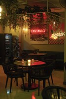 Interior de un restaurante tenuemente iluminado con mesas, sillas, plantas colgantes y un letrero de neón.