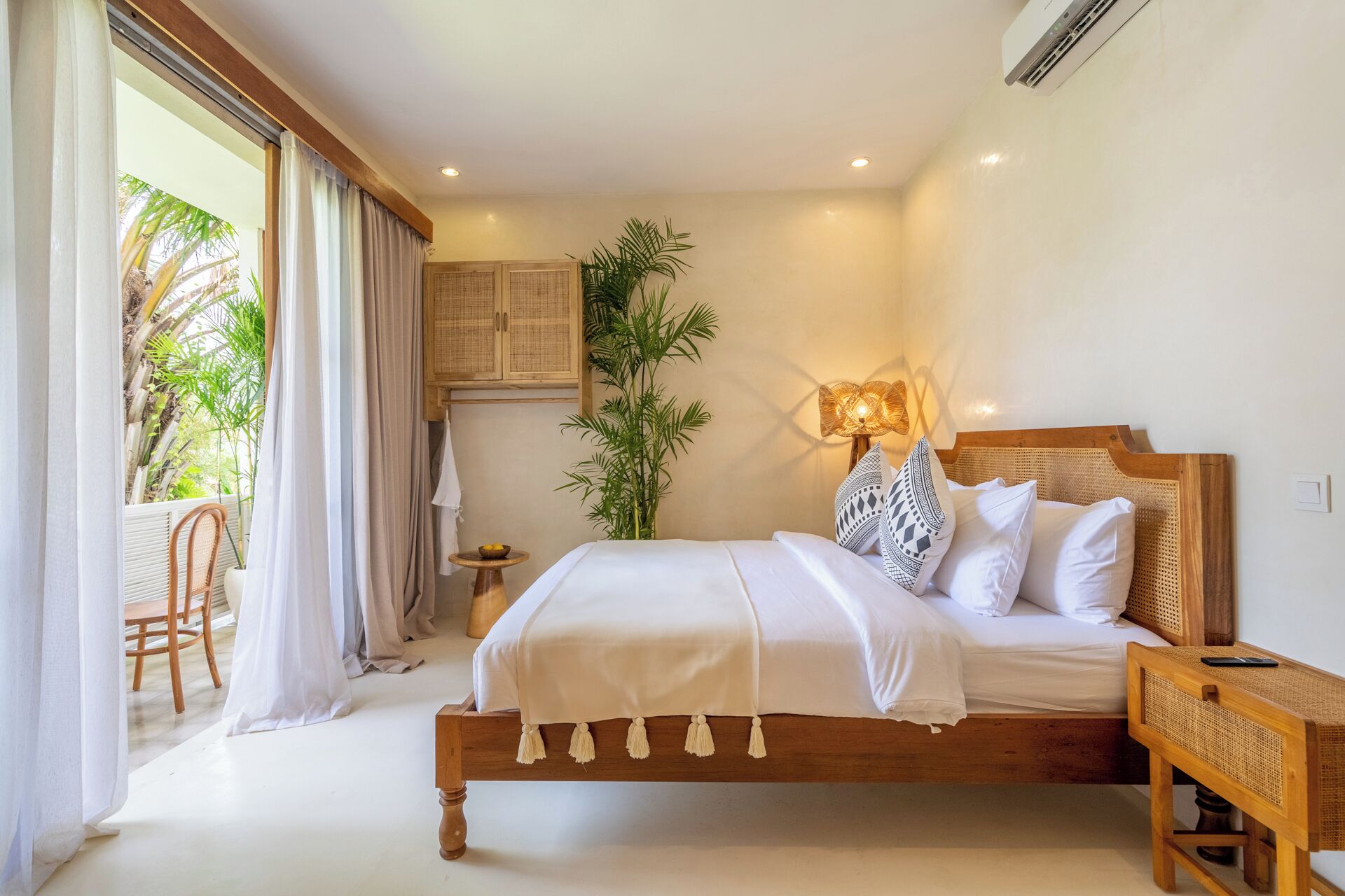 A bedroom with a wooden bed, white bedding, a bedside lamp, and a view of a balcony with plants.