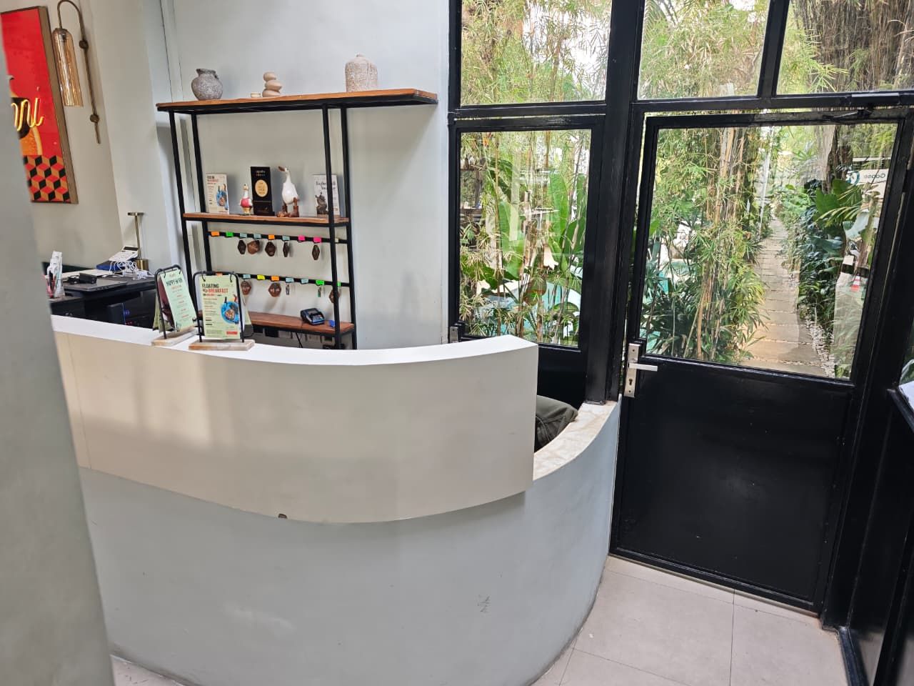 A reception desk with a curved design, a shelf with decorative items, and a view of greenery through a large window.