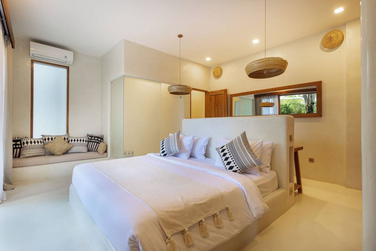 Een slaapkamer met een groot bed, een inbouwkbank met kussens, een spiegel en een raam met jaloezieën.