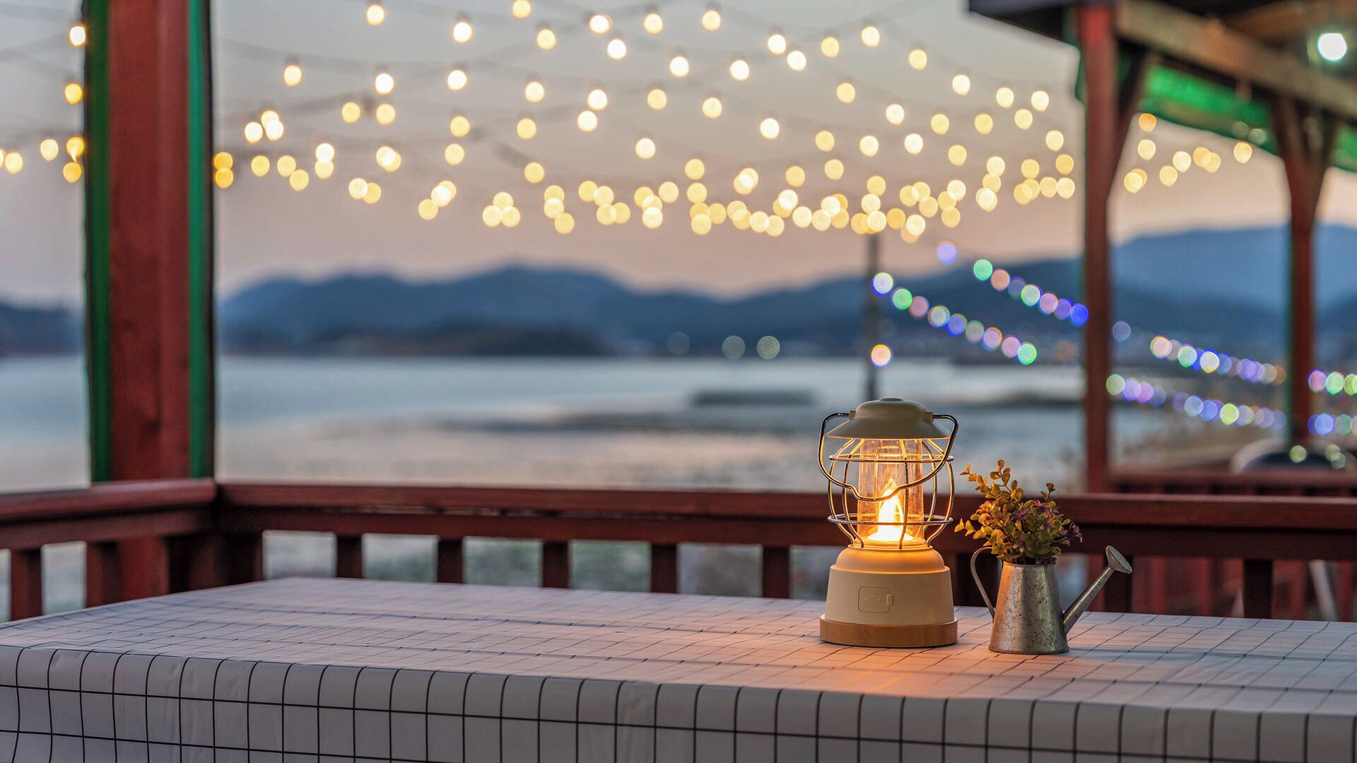Una mesa con un farolillo y una jarra metálica, frente a una vista de agua y montañas, con luces colgantes.
