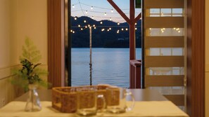 A room with a view of a lake and mountains, a table with a vase and baskets, and a wooden structure with string lights. - Namhae Bada Yanolja Pension (Namhae)