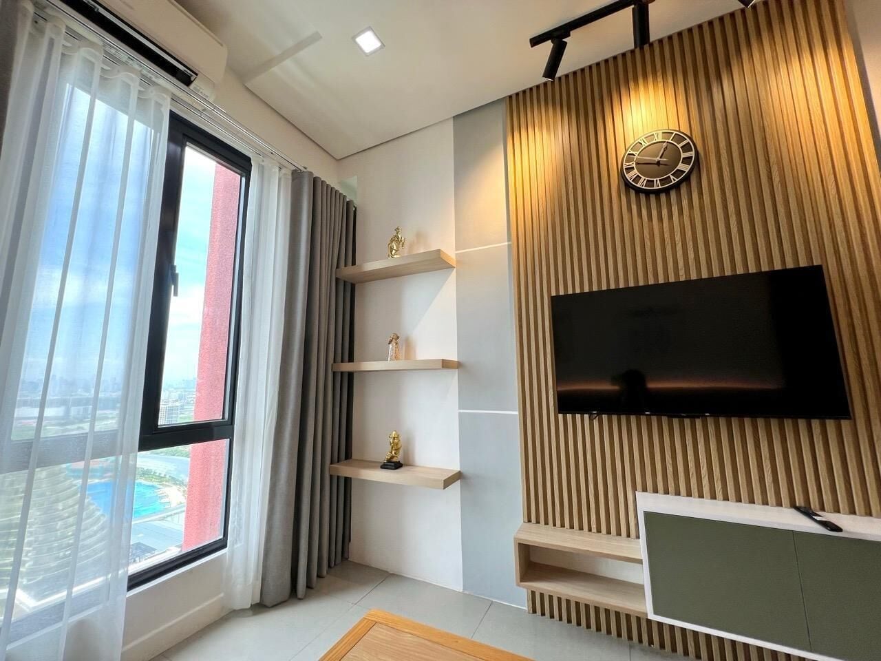 A modern living room with a large window, a flat-screen TV mounted on a wooden wall, and built-in shelves.