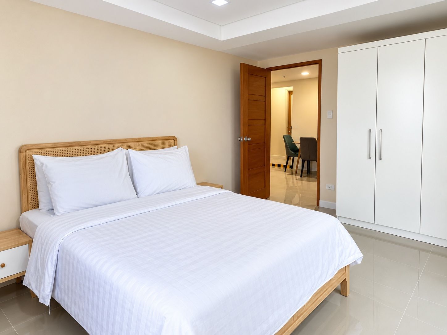 A bedroom with a bed, a wooden nightstand, a wardrobe, and a door leading to a dining area.