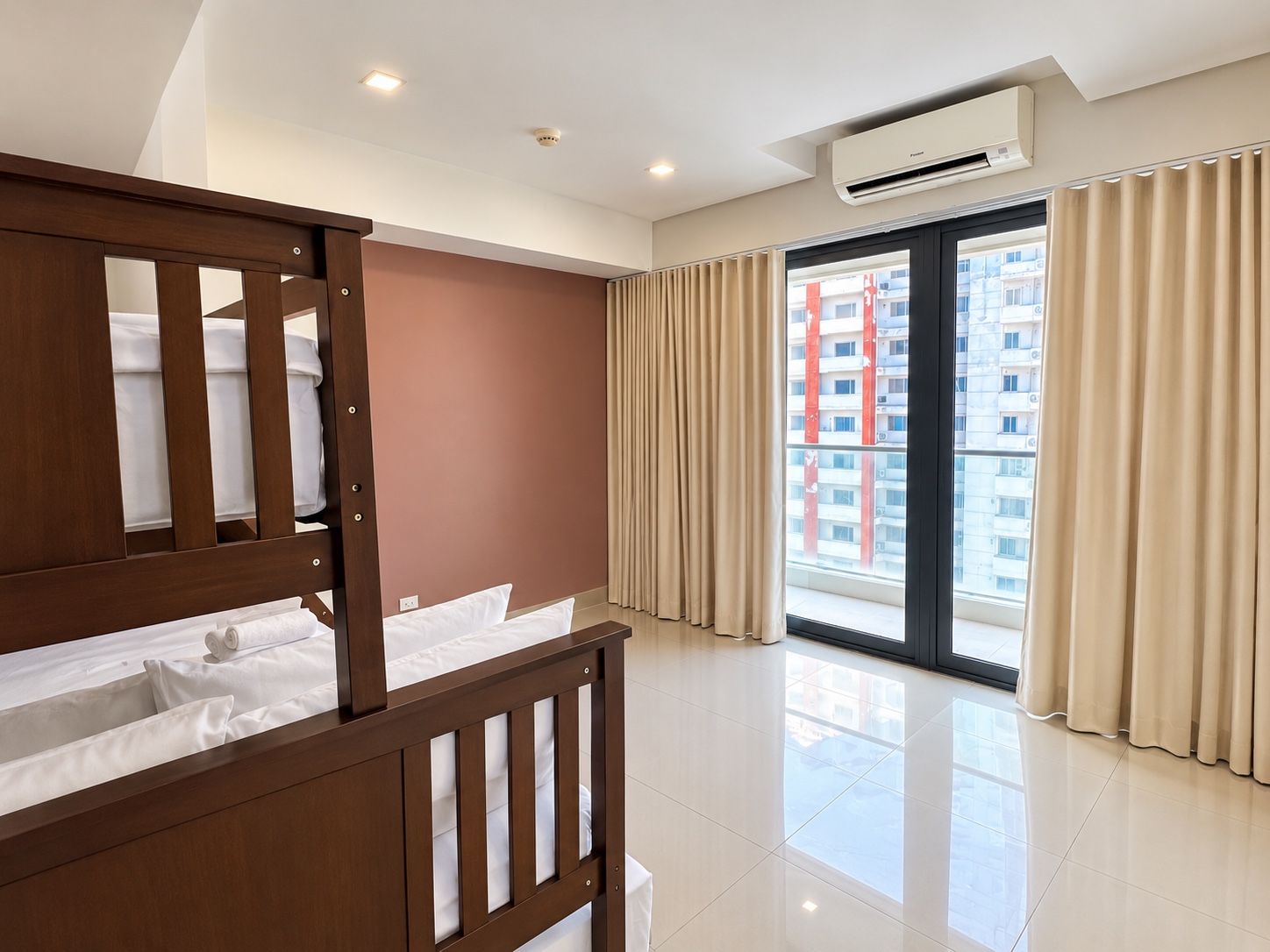 A hotel room with a wooden bed, a large window with curtains, a ceiling air conditioner, and a view of a building outside.