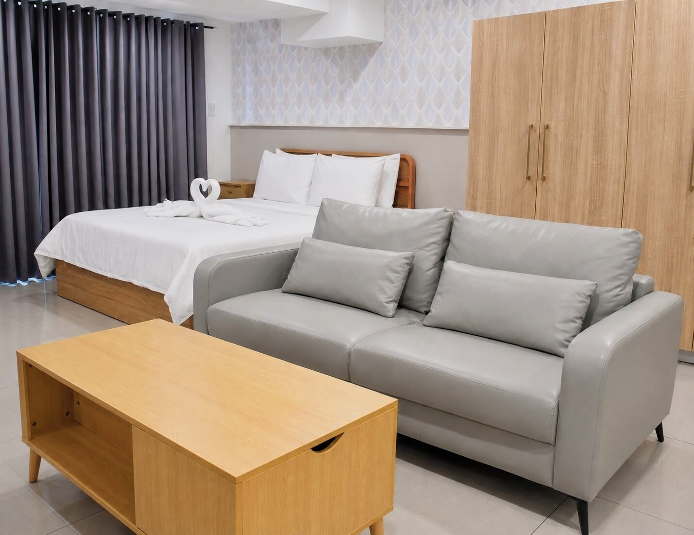 A modern hotel room with a bed, a grey sofa, a wooden coffee table, and a wardrobe.