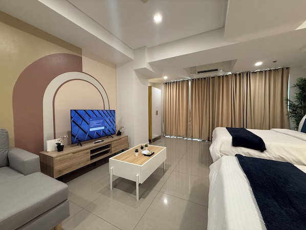 A modern hotel room with a flat-screen TV on a wooden console, a white coffee table, a bed with a dark throw, and a large window with curtains. - Sapphire Seaview Park (Parañaque)