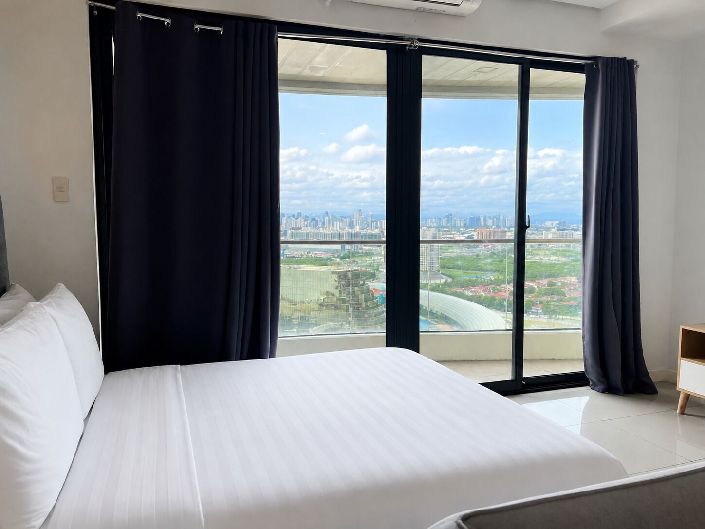 A hotel room with a large bed, dark curtains, and a view of a cityscape through a sliding glass door.