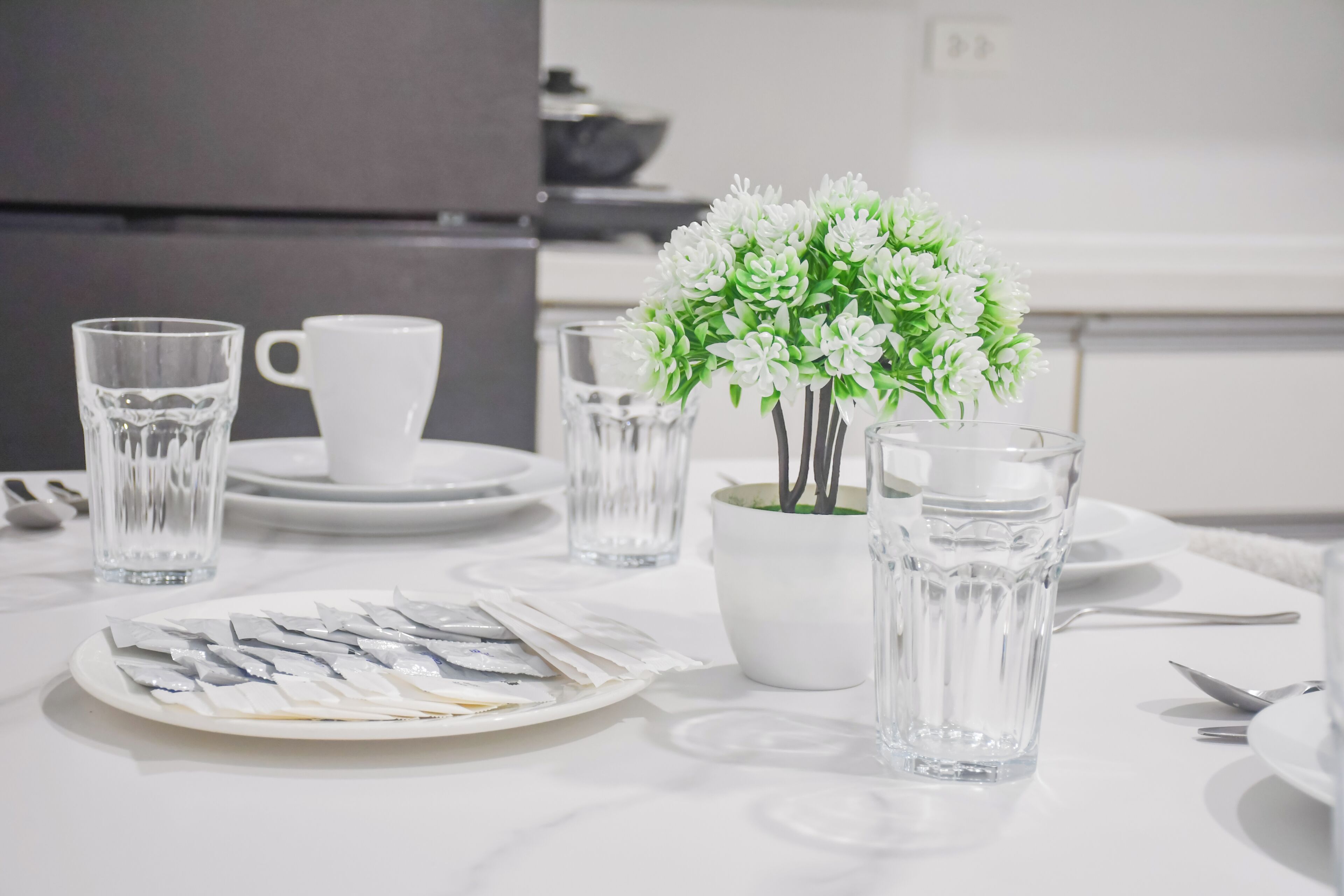 Uma mesa com um guardanapo de papel branco, dois copos de vidro transparentes, uma chávena branca e um vaso com flores brancas.