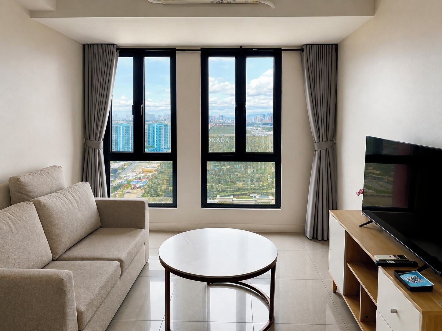 A modern living room with a sofa, a round table, a flat-screen TV, and a city view through large windows.