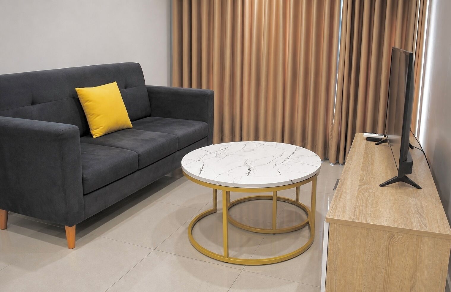 Una sala de estar moderna con un sofá gris, una almohada amarilla, una mesa de centro de mármol y un televisor sobre un mueble de madera.