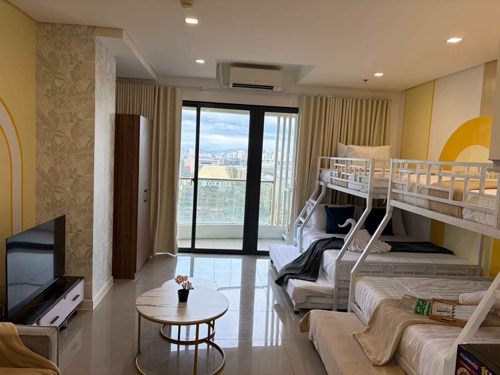 A hotel room with a TV, a small round table, a sofa, a bunk bed, and a balcony with a city view.