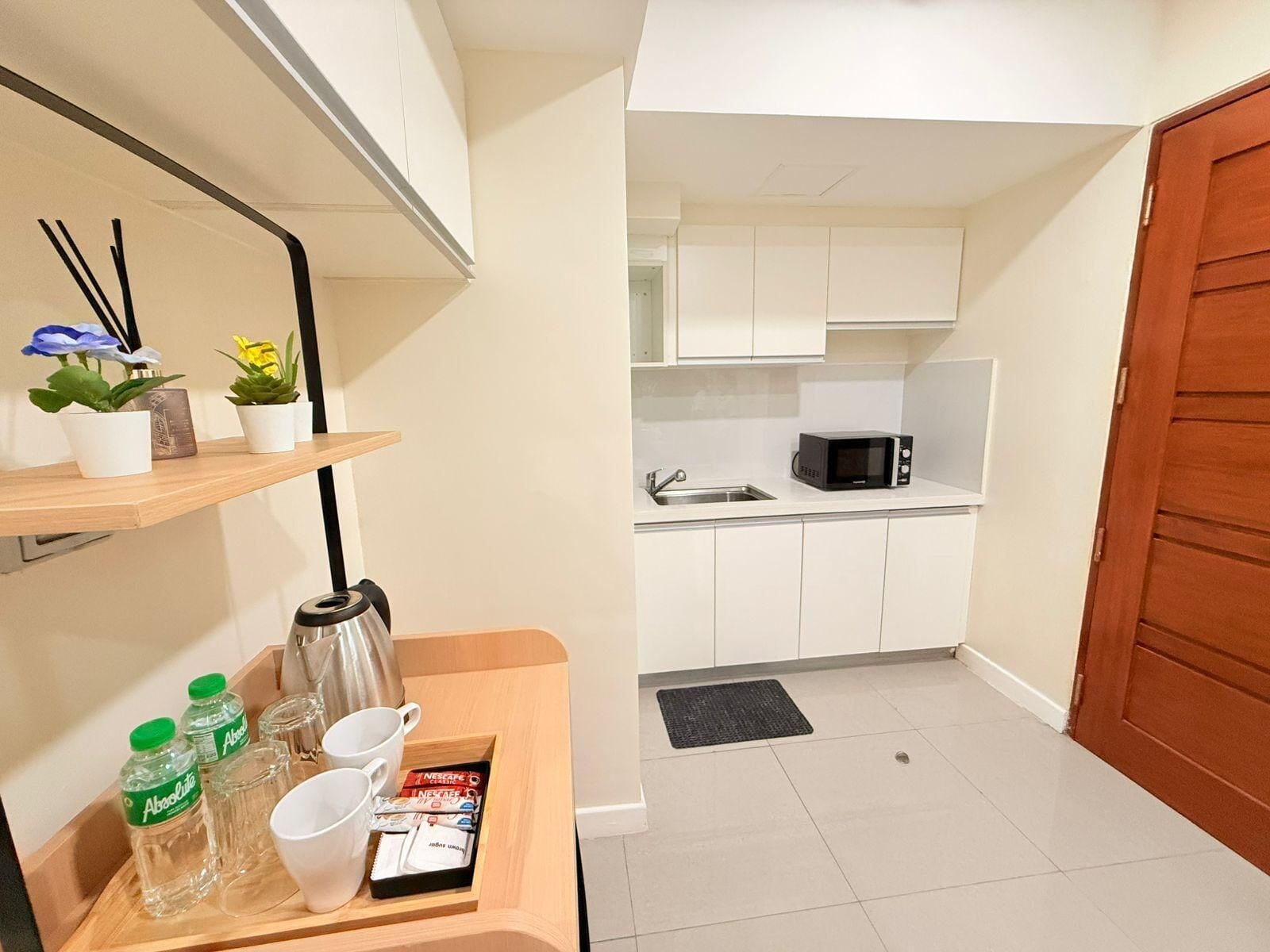 A compact kitchen with white cabinets, a microwave, and a sink. There is a small table with a kettle, cups, and a tray with coffee packets.
