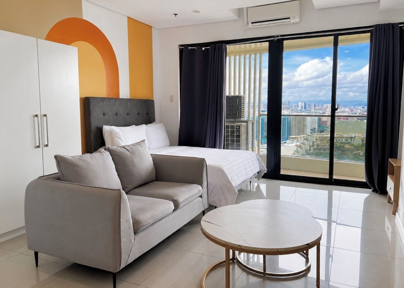 A modern hotel room with a bed, a sofa, a small table, and a city view through large windows.