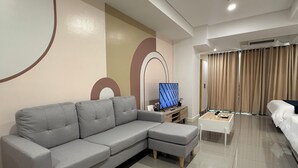 A modern living room with a grey sectional sofa, a flat-screen TV on a wooden stand, a white coffee table, and a bed with beige curtains. - Sapphire Seaview Park (Parañaque)
