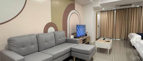 A modern living room with a grey sectional sofa, a flat-screen TV on a wooden stand, a white coffee table, and a bed with beige curtains.