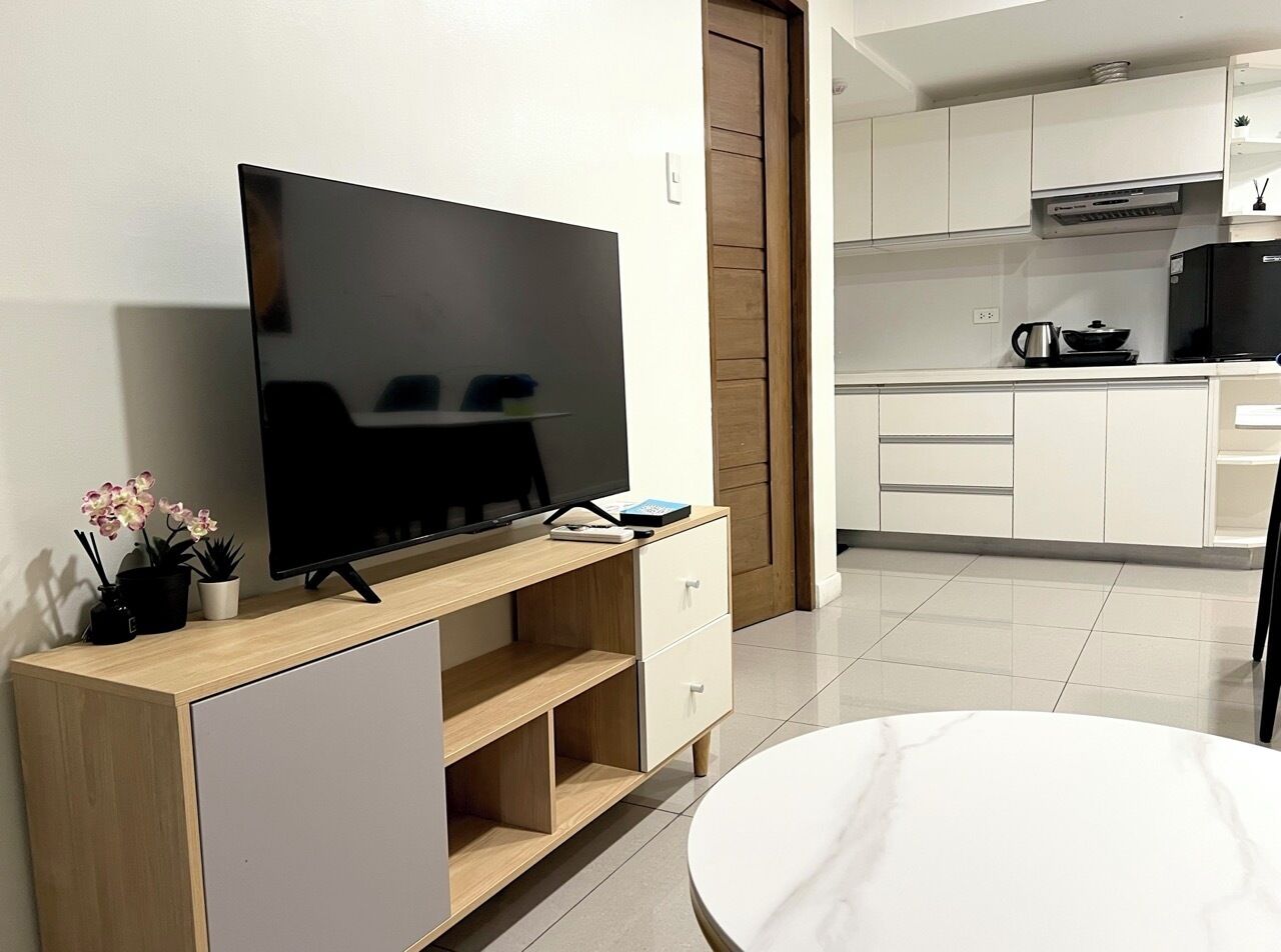 Un salon moderne avec un téléviseur à écran plat posé sur une console en bois, une table à manger blanche et un coin cuisine à l’arrière-plan.