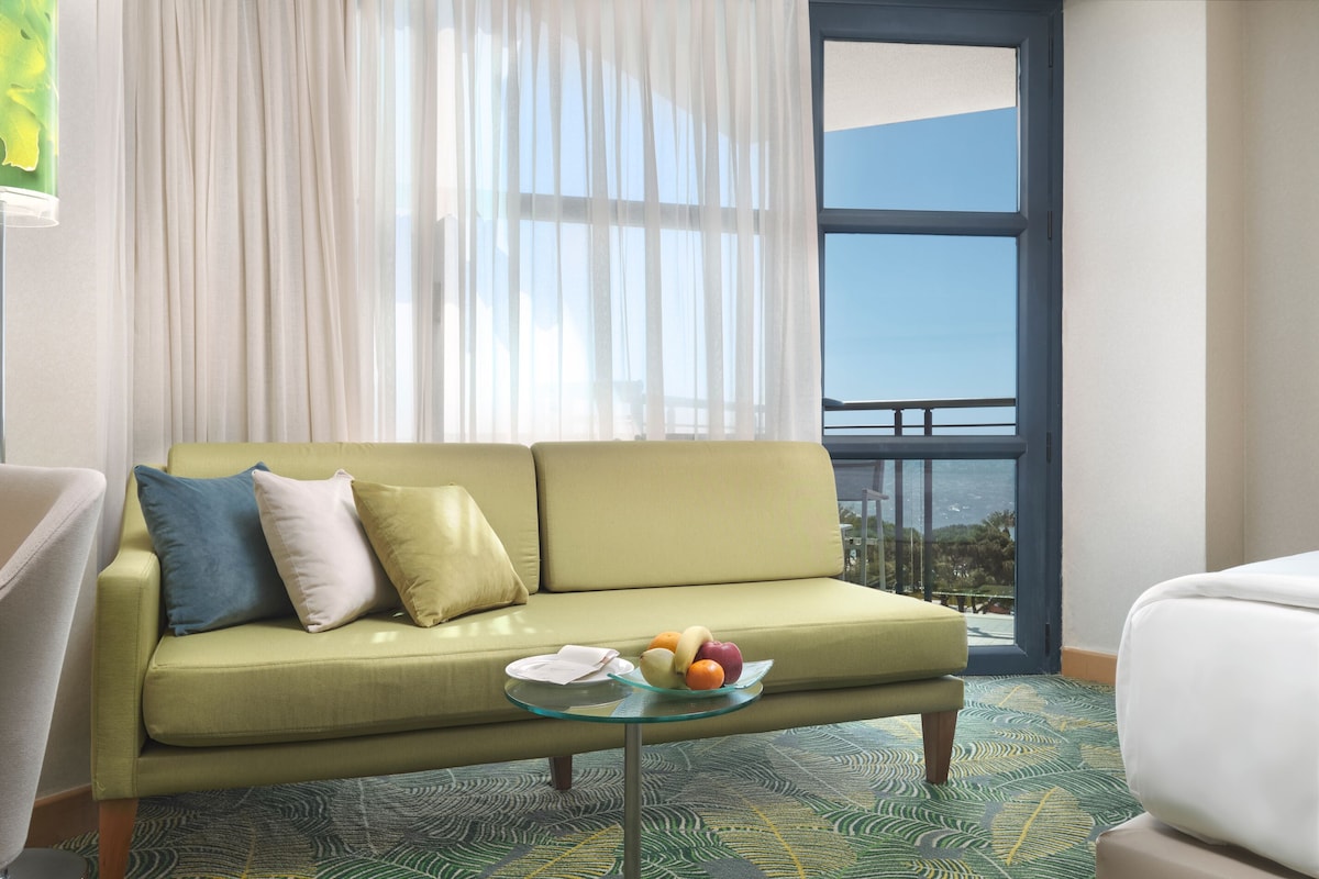 A green sofa with cushions, a glass coffee table with fruit, and a view of the outdoors through a window.