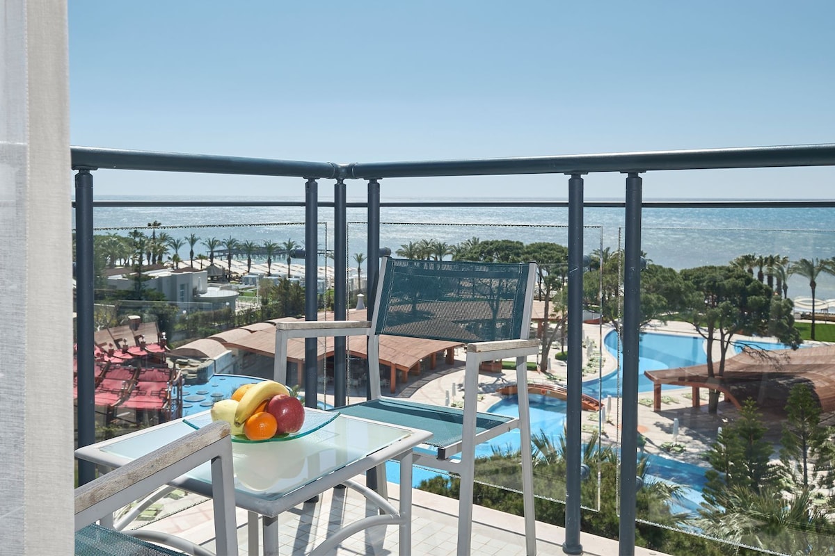 A balcony with a glass table and chairs, overlooking a pool area with a view of the beach and palm trees.