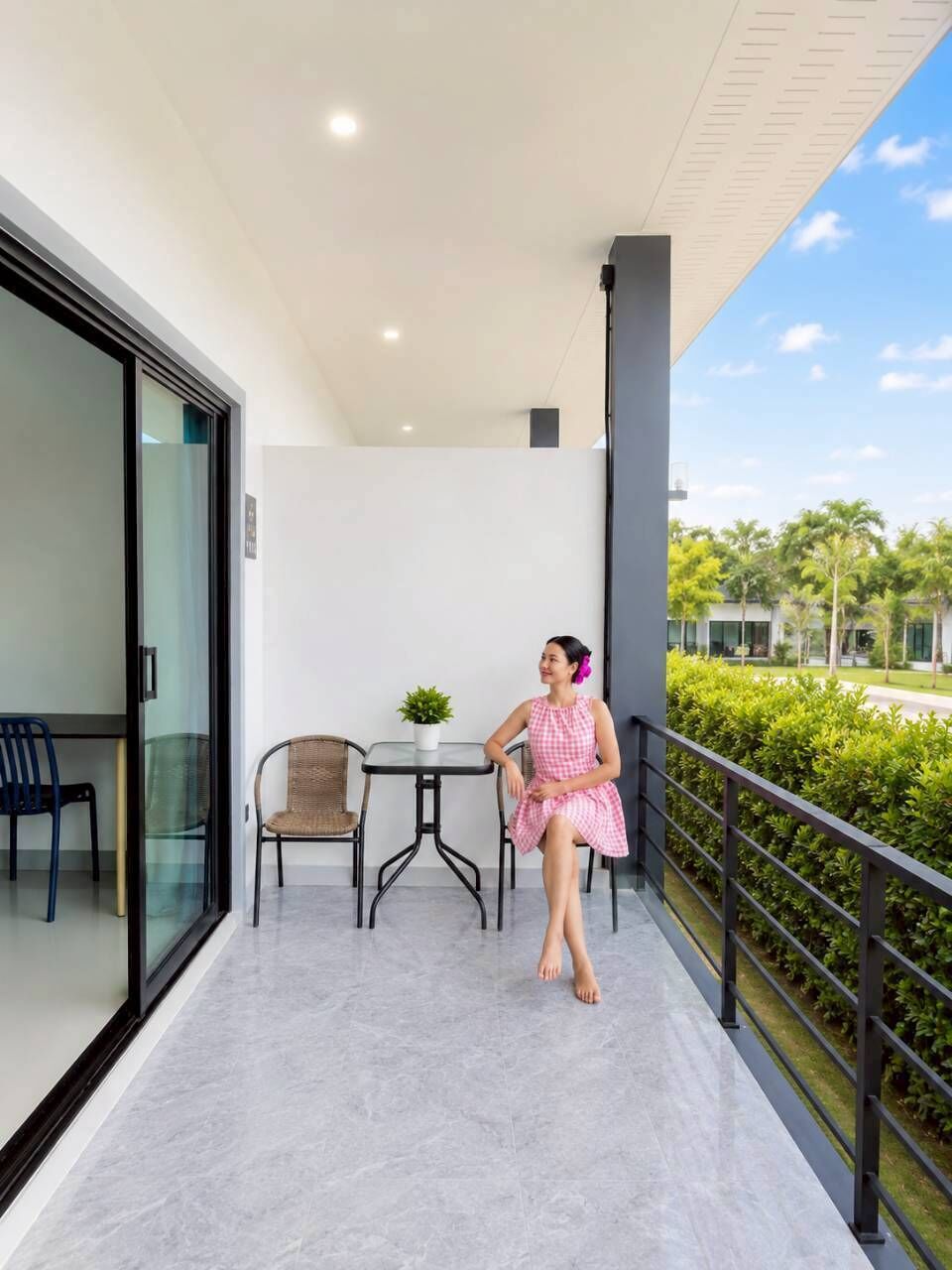 A balcony with a glass door, a small table with a plant, a wicker chair, and a person in a pink dress.