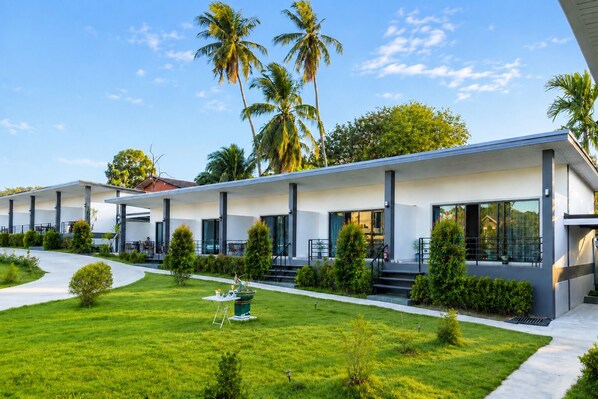 A modern hotel with a covered outdoor seating area, a well-maintained lawn, and palm trees in the background.