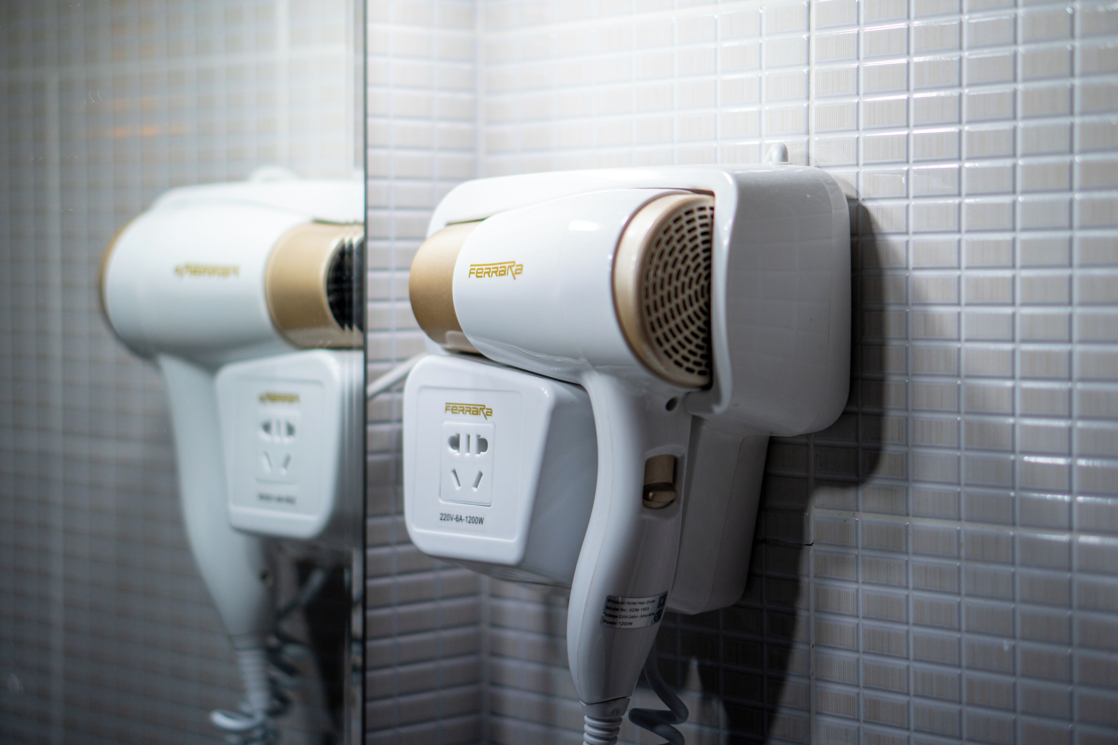 A hair dryer mounted on a tiled wall, with a visible power outlet and a cord.