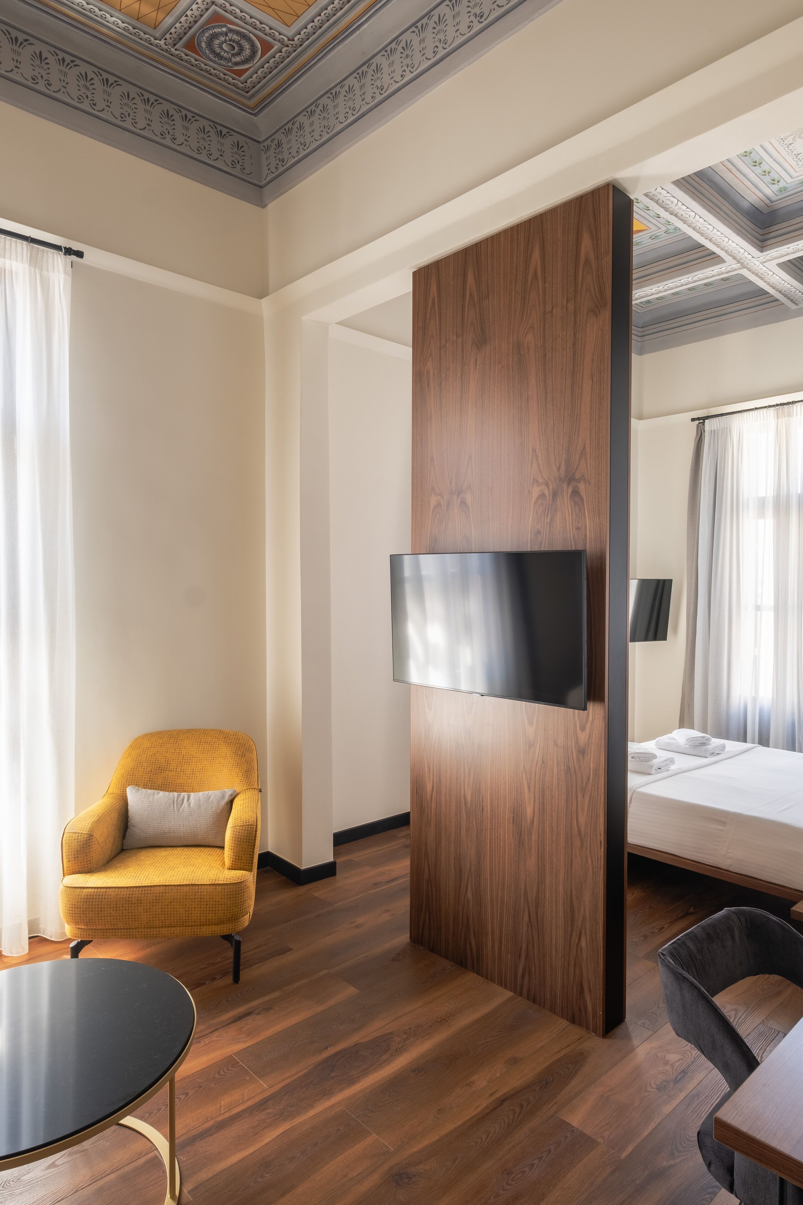 Habitación de hotel con un panel de madera, un televisor de pantalla plana, una cama, un sillón amarillo, una mesa redonda y un techo con diseños decorativos.