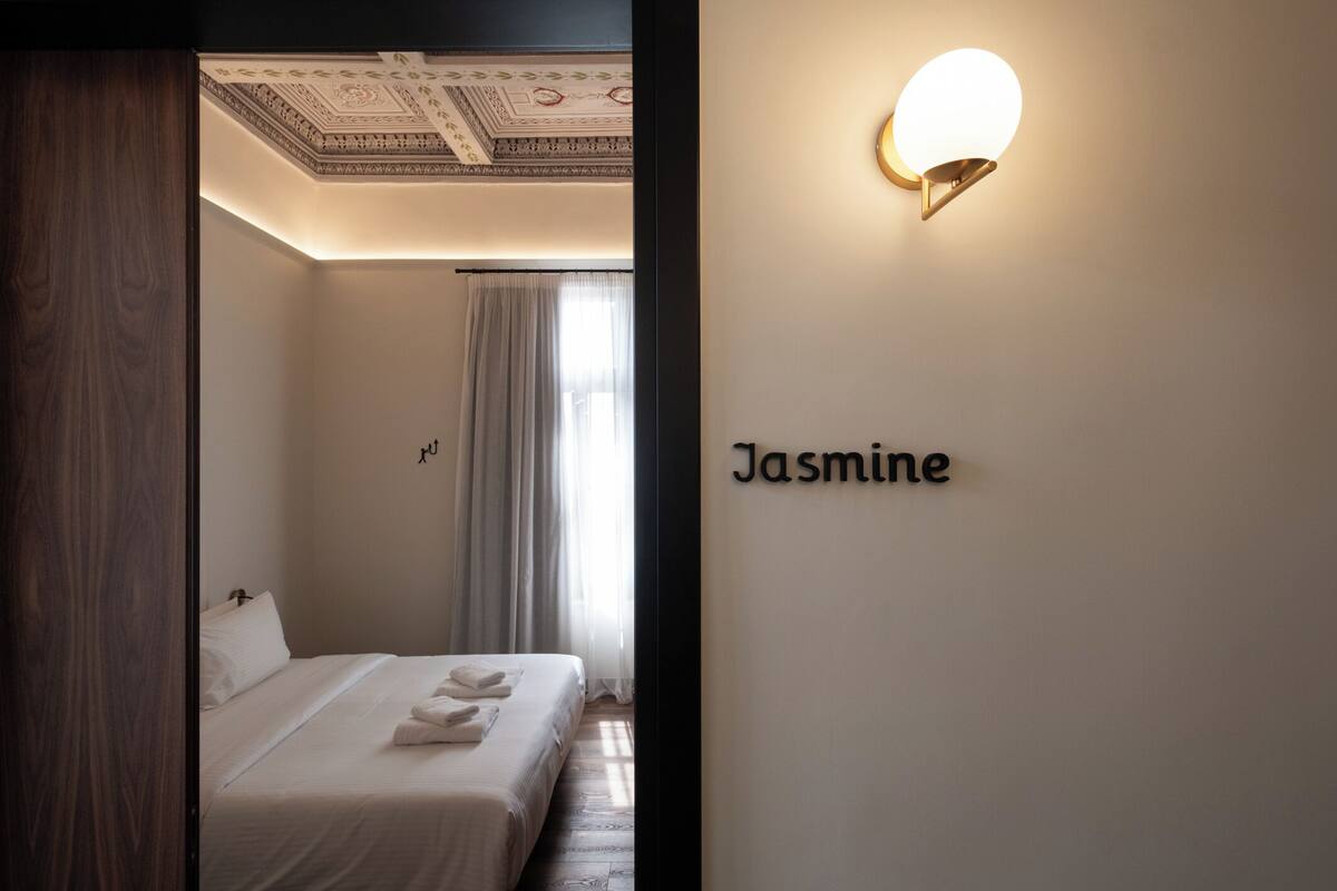 Quarto de hotel com uma cama bem arrumada, luminária fixada na parede e uma placa com o nome 'Jasmine'.