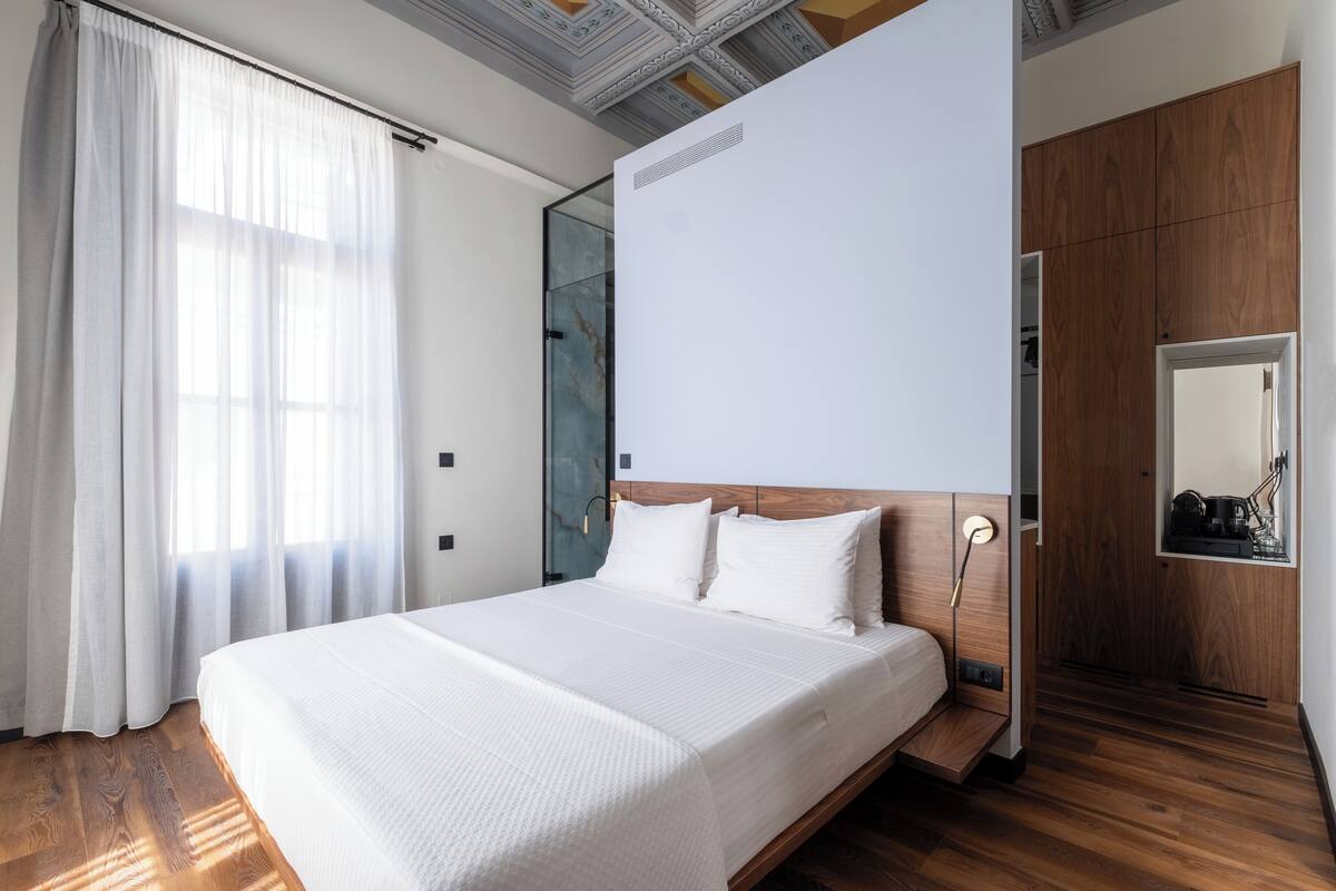Quarto de hotel moderno com uma cama grande, piso de madeira, peseira de madeira e uma janela ampla com cortinas leves.