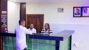 A reception desk with a wooden door in the background, framed pictures on the wall, and a sign with text. - New Eastern Comfort Hotel Umuahia (Umuahia)