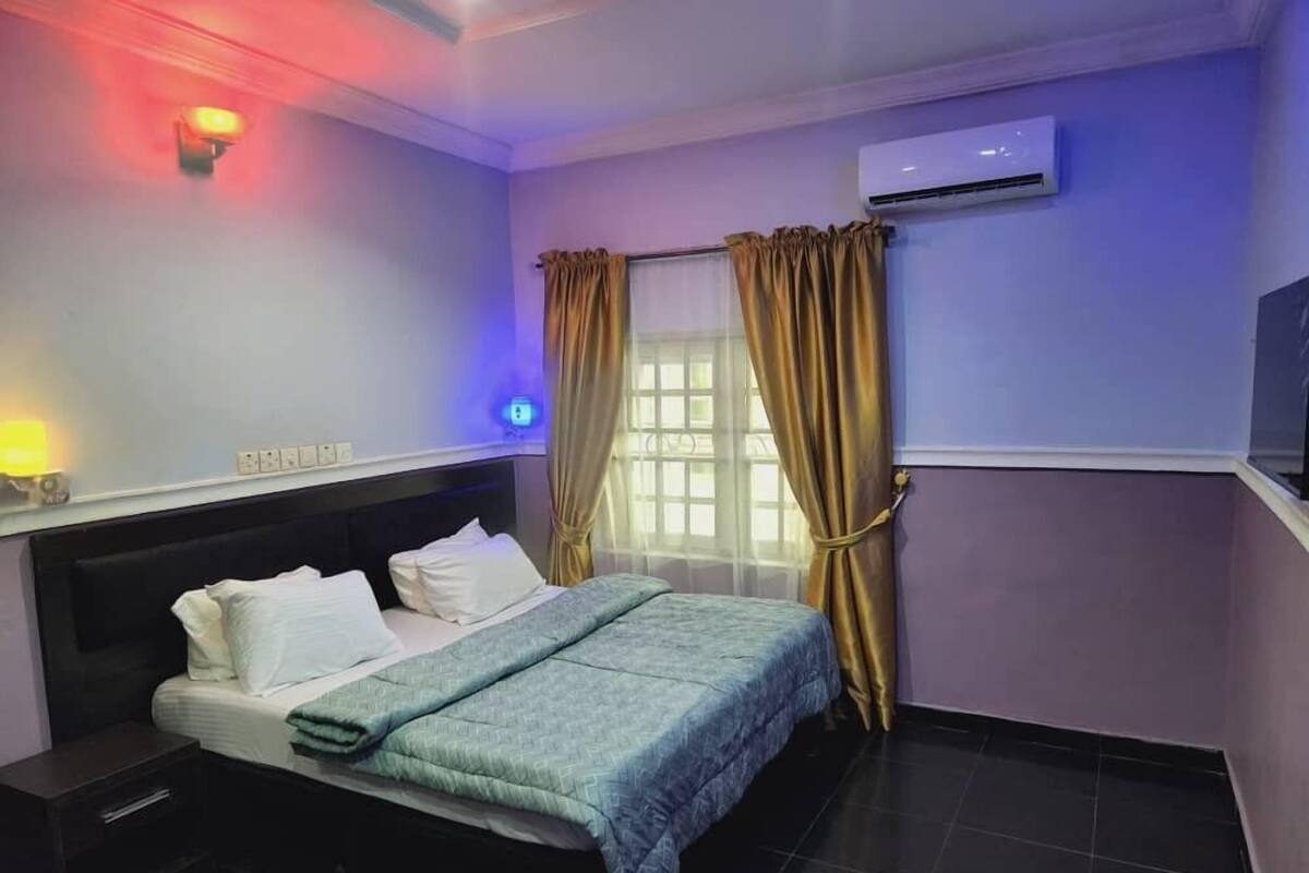 A hotel room with a bed, bedside table, air conditioning unit, and curtains.